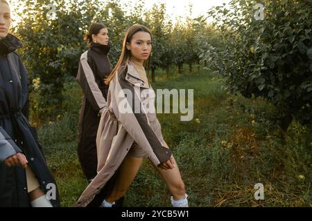 Drei stylische Mädchen in Regenmänteln in einem Apfelgarten. Eine Modenschau im Garten bei Sonnenuntergang. Stockfoto