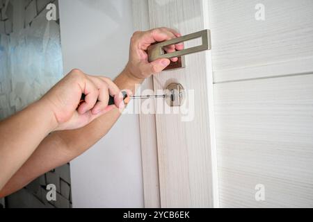 Der Handwerker installiert einen neuen Türgriff an einer weißen Tür mit einem Schraubendreher. Stockfoto