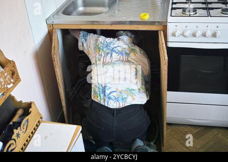 Ein Mann auf den Knien, der Wasserarmaturen in einer Küche repariert, Innenaufnahme Stockfoto