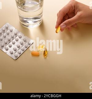 Weiße und gelbe Pillen, Vitamine oder Nahrungsergänzungsmittel liegen auf dem Tisch neben einem Glas Wasser, in der Hand befindet sich eine Kapsel mit Omega 3, Kopierraum Stockfoto