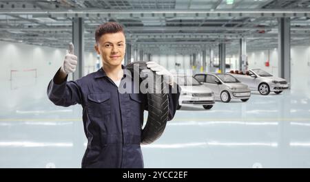 Ein junger Mechaniker, der einen Motorradreifen trägt und in einer Autogarage die Daumen hoch zeigt Stockfoto