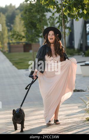 Eine Frau in Kleid und Hut mit ihrem französischen Bulldogge-Hund auf einem Spaziergang durch die Stadt. Stockfoto