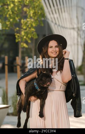 Eine Frau in Kleid und Hut hält ihren geliebten französischen Bulldogge-Hund vor dem Hintergrund der Stadt in den Armen Stockfoto