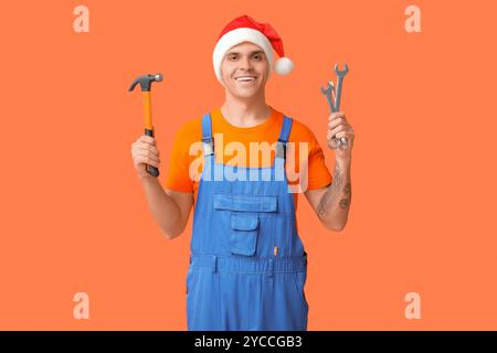 Männlicher Klempner im Weihnachtsmann-Hut mit Hammer und Schraubenschlüsseln auf orangem Hintergrund Stockfoto