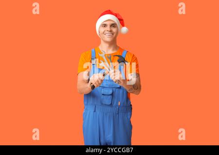Männlicher Klempner im Weihnachtsmann-Hut mit Hammer und Schraubenschlüsseln auf orangem Hintergrund Stockfoto