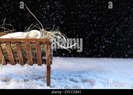 Dummy von Baby und Holzkrippe mit Heu auf Schnee vor schwarzem Hintergrund. Das Konzept der Weihnachtsgeschichte Stockfoto