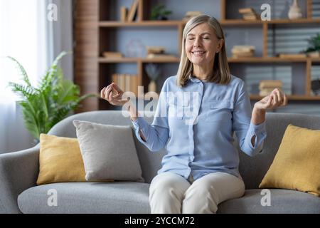 Reife Frau meditiert auf der Wohnzimmercouch mit geschlossenen Augen, um Ruhe und Entspannung zu genießen. Die ruhige Atmosphäre zu Hause fördert die gelassene Achtsamkeit Stockfoto