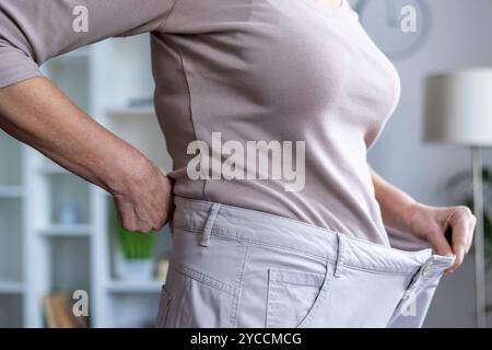 Die Frau zeigt deutliche Erfolge bei der Gewichtsreduktion, indem sie den Bund der losen Hose hält. Feierlicher Moment unterstreicht Engagement für gesundes Leben und Fitness Leistungen. Stockfoto