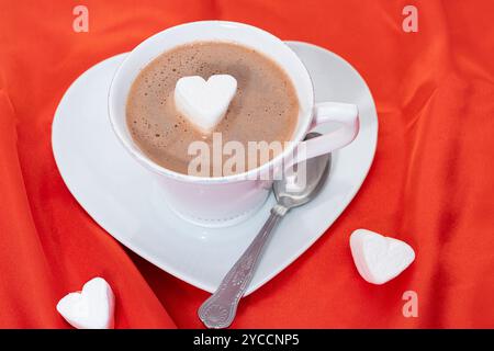 Eine Tasse heiße Schokolade mit einem herzförmigen Marshmallow - roter Satin Hintergrund Stockfoto