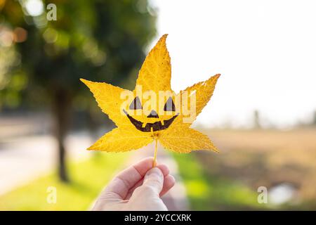 Die Hand hält ein gelbes flüssiges Blatt mit der Zeichnung eines halloween Kürbisgesichts drauf. Herbsthintergrund mit Kopierraum. Stockfoto