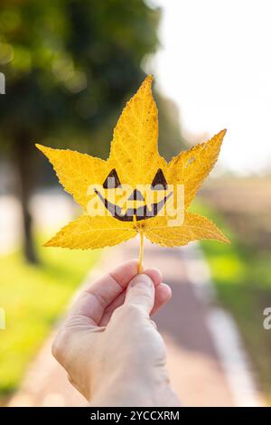 Die Hand hält ein gelbes flüssiges Blatt mit der Zeichnung eines halloween Kürbisgesichts drauf. Herbsthintergrund mit Kopierraum. Vertikale Aufnahme. Stockfoto