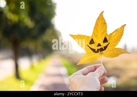 Die Hand hält ein gelbes flüssiges Blatt mit der Zeichnung eines halloween Kürbisgesichts drauf. Herbsthintergrund mit Kopierraum. Stockfoto