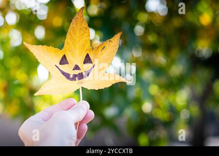 Die Hand hält ein gelbes flüssiges Blatt mit der Zeichnung eines halloween Kürbisgesichts drauf. Herbsthintergrund mit Kopierraum. Stockfoto