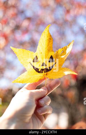 Die Hand hält ein gelbes flüssiges Blatt mit der Zeichnung eines halloween Kürbisgesichts drauf. Herbsthintergrund mit Kopierraum. Vertikale Aufnahme. Stockfoto