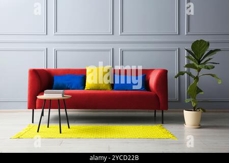 Stilvolles rotes Sofa mit Kissen, grüner Pflanze und Tisch im Zimmer Stockfoto