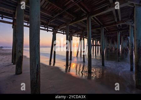 Sonnenlicht filtert durch die hölzernen Stützen eines Piers und wirft Reflexionen auf den nassen Sand darunter. Die ruhige Meereskulisse verstärkt den friedlichen geldautomaten Stockfoto