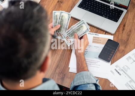 Grauhaariger Mann zählt hundert Dollar-Banknoten über Stromrechnungen, die mit Laptop am Tisch sitzen Stockfoto