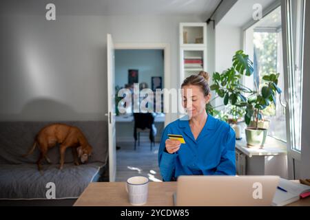 Eine Frau mittleren Alters, die eine Kreditkarte hält, die am Tisch sitzt und mit Laptop positivem Online-Shopping teilnimmt Stockfoto
