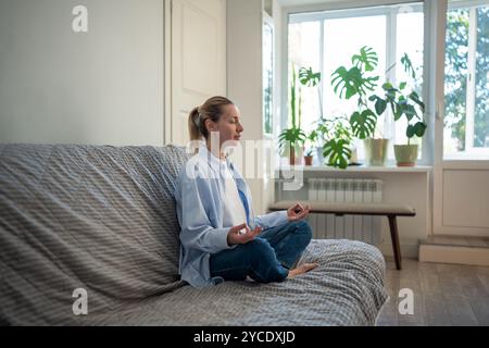 Eine ruhige Frau mit geschlossenen Augen übt achtsames Atmen auf der Couch in Padmasana Lotus Pose Stockfoto