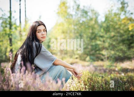 Eine junge Frau sitzt ruhig auf dem Waldboden, ihr langes Haar zieht sich über den Rücken. Sie erscheint tief in Gedanken, umgeben von blühenden Wildblumen Stockfoto