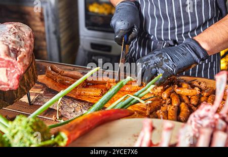 Ein Koch in schwarzen Handschuhen schneidet Würstchen, umgeben von frischem Gemüse und Fleisch auf einem Holzbrett. Stockfoto