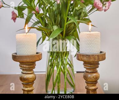 Ein Paar Kerzen auf eleganten Holzhaltern neben einer Vase mit frischen rosa Blumen, die eine gemütliche Atmosphäre schaffen. Stockfoto