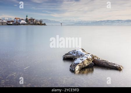 Holzstamm und malerische Kirche St. George am Seeufer vor dem schneebedeckten Pfaender an einem klaren Wintertag, Wasserschloss, Lake Const Stockfoto