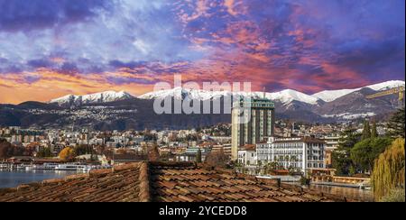 Sonnenuntergang in Lugano und schneebedeckte Berge und Wald dahinter. Stadtbild und Kornett unten links und Herbstblattfarbe der Bäume am sonnigen Tag von Anfang des Jahres Stockfoto