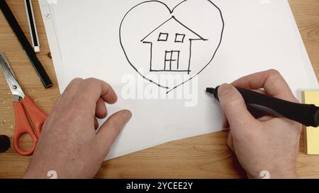 Die Hand eines Innenarchitekten zeichnet ein Haus in einem Herzen auf einem Blatt Papier, das ideale Filmmaterial für Themen wie Zuhause, Familie und neues Haus Stockfoto