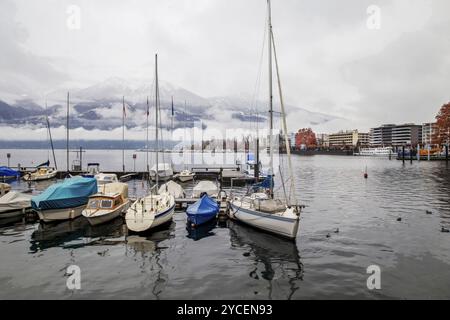 Nebelnde Naturszene und geschütztes Segelboot in kleinem Hafen und Pier im Winter Wasser und orange Lärchen in der Ferne und in der Stadt Locarno Stockfoto