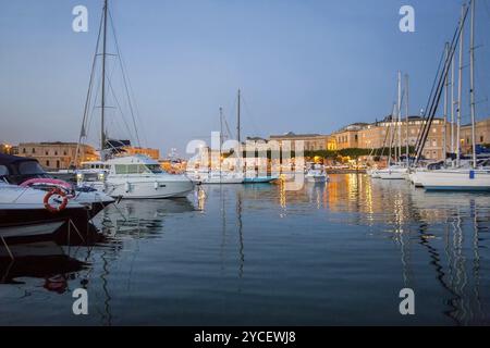 Kleine Segelboote auf den Parkplätzen im Hafen von Siracuse. Hinter der Altstadt von Sirakus und den elektrischen Lichtern der Stadt (Straßenluken) und einer Gasse mit Kiefern! Stockfoto