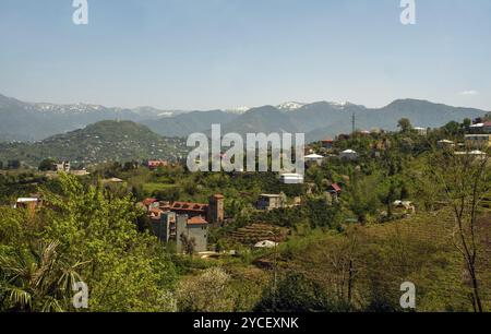 Idyllisches Panorama auf den Hügeln in der Nähe von Batumi mit kleinen Häusern (mit Gärten und Obstgärten) und schneebedeckten Hügeln Ende April. Landwirtschaftliche Terrassenfelder Stockfoto