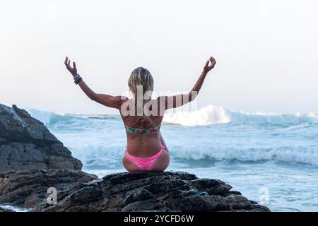 Blonde Frau mit lockeren Haaren in einem Bikini, die auf einer Klippe vor dem Meer sitzt, das vom Wasser der Wellen bespritzt wird Stockfoto