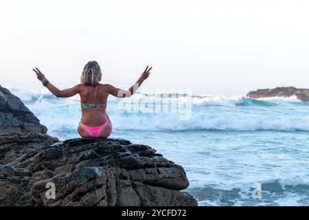 Blonde Frau mit lockeren Haaren in einem Bikini, die auf einer Klippe vor dem Meer sitzt, das vom Wasser der Wellen bespritzt wird Stockfoto