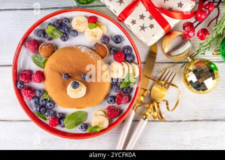 Lustige und süße Weihnachts-Teddybären-Pfannkuchen auf einem rot-weißen Teller, dekoriert mit frischen Beeren, Obst und Puderzucker, Kinderweihnachtsfrühstück Stockfoto