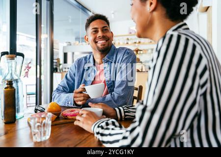 Wunderschönes junges gemischtes Paar, das sich in einer Bar-Cafeteria zusammenhält und sitzt - ziemlich fröhliche afroamerikanische Freunde, die sich in einem Gebäck-Bäckerei-Kaffee entspannen Stockfoto