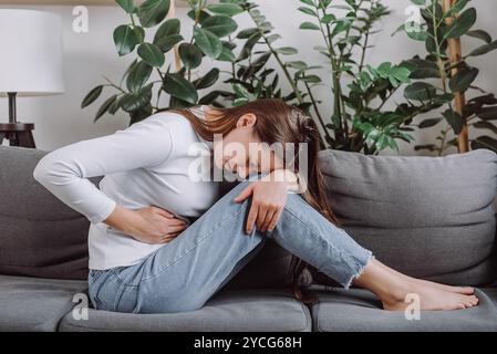 Verärgerte Frau, die auf dem Sofa sitzt und unter Verdauungsstörungen leidet, Blähungen und Bauchproblemen. Kranke junge Frau mit Schmerzen, die Bauchgefühl h hält Stockfoto