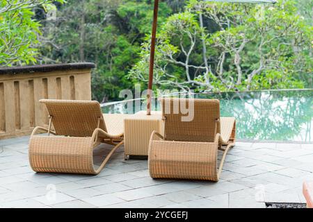 Zwei Rattanliegestühle mit Sonnenschirm auf einer Terrasse mit Blick auf eine üppige grüne Landschaft. Die Stühle befinden sich in der Nähe eines Glasgeländers und bieten ein V Stockfoto