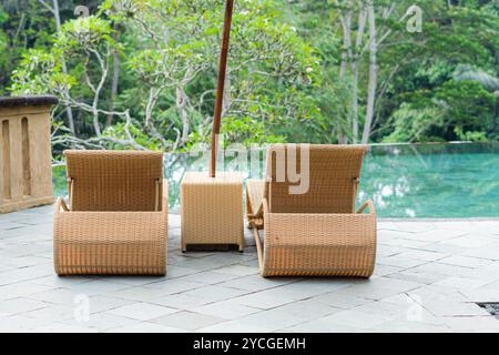 Zwei Rattanliegestühle mit Sonnenschirm auf einer Terrasse mit Blick auf eine üppige grüne Landschaft. Die Stühle befinden sich in der Nähe eines Glasgeländers und bieten ein V Stockfoto