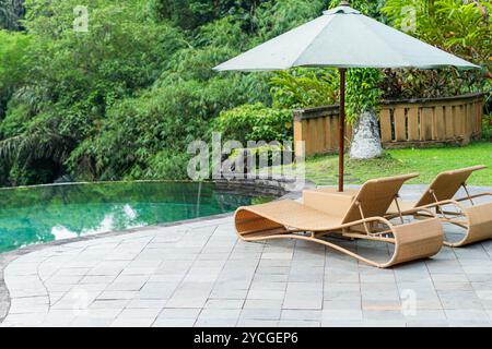 Zwei Rattanliegestühle mit Sonnenschirm auf einer Terrasse mit Blick auf eine üppige grüne Landschaft. Die Stühle befinden sich in der Nähe eines Glasgeländers und bieten ein V Stockfoto