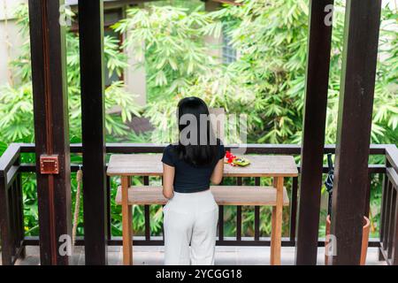 Eine Frau steht an einem Holztisch in einer üppig grünen Umgebung im Freien, umgeben von tropischen Pflanzen. Sie trägt ein schwarzes Oberteil und eine weiße Hose Stockfoto