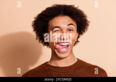 Junger Mann mit fröhlichem Ausdruck, der zwinkert und die Zunge vor beigefarbenem Hintergrund in lässigem Pullover ausstreckt Stockfoto