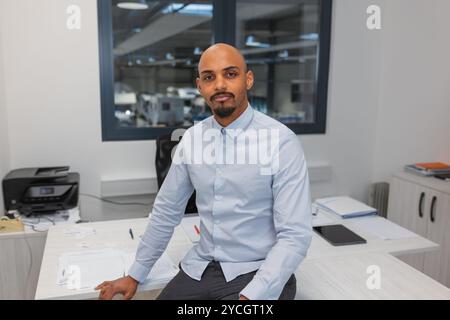 Porträt eines selbstbewussten jungen afroamerikanischen Geschäftsmannes, der ein elegantes weißes Hemd trägt und sich lässig in einem Firmenbüro posiert. Stockfoto