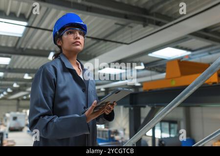 Eine junge Ingenieurin mit blauem Helm und Arbeitsmantel posiert in einer Fabrik mit einem Tablet, Porträt aufgenommen. Berufsfrauen im Industriekonzept Stockfoto