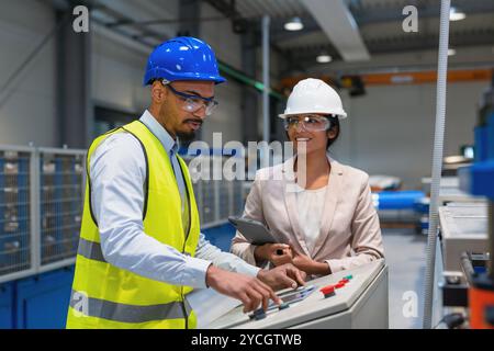Die indische Betriebsleiterin trainiert einen afroamerikanischen männlichen Mitarbeiter in einem blauen Helm und einer gelben Weste, um Maschinen zu bedienen. Stockfoto