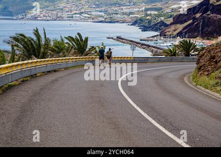 Zwei Wanderer, die auf einer gekrümmten Straße mit Blick auf die Küstenstadt und den Yachthafen in der Nähe von Vereda da Ponta de São Lourenco spazieren Stockfoto