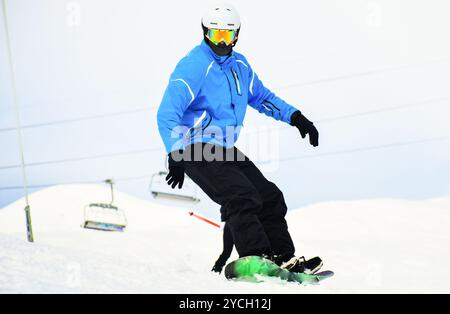 Nahaufnahme lässiger Snowboardfahrer im blau-schwarzen Outfit blickt auf die Kamera isoliert in weißem, verschneiten Hintergrund. Winterurlaub mit Sportbergen Stockfoto