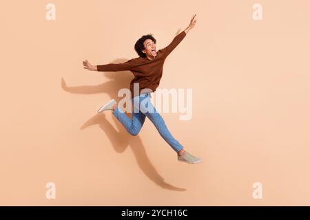 Junger Mann, der fröhlich in lässigem Outfit vor beigefarbenem Hintergrund für Urlaubsangebote springt Stockfoto