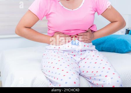 Nahaufnahme einer jungen Frau, die ihren Bauch drückt, während sie auf einem Bett sitzt und an Menstruationskrämpfen leidet. Sie ist in einem gut beleuchteten Raum Stockfoto