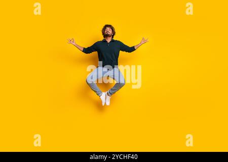 Junger Mann in schwarzem Hemd und Jeans, der an einem zwanglosen freitag fröhlich vor einem hellgelben Hintergrund springt Stockfoto